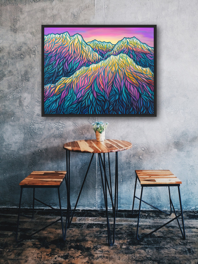 Framed colorful mountain landscape art on a textured wall above a small wooden table and stools.