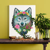 Art print of a wolf with floral crown on a wooden table against a yellow wall.