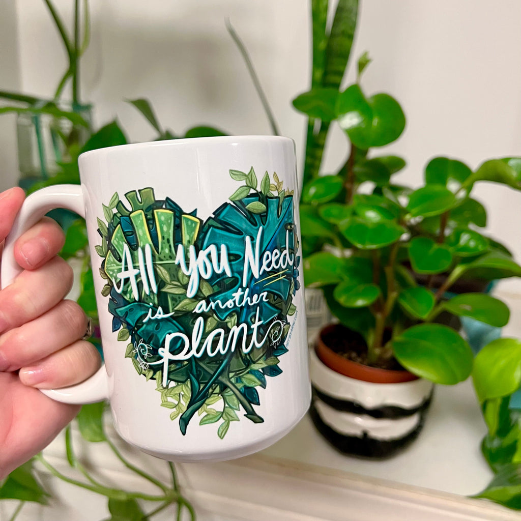 hand holding White coffee mug with plants in the background and on the cup is Monstera Leafs and house plants assorted into a hear shape with white text that says "All You Need Is Another Plant" 